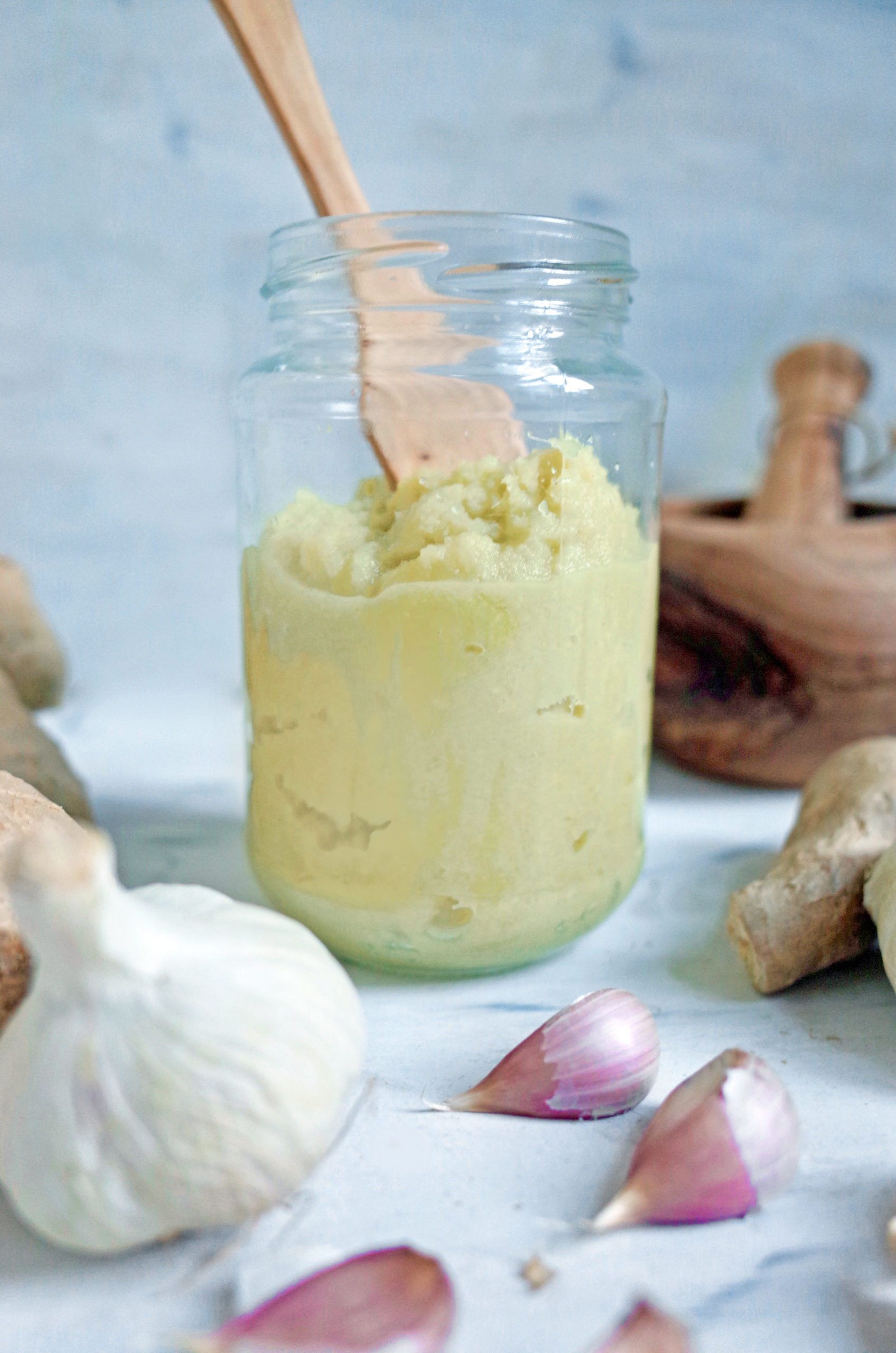 A jar of homemade ginger garlic paste with a wooden spoon and garlic bulbs
