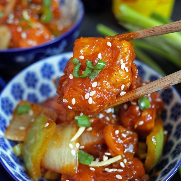 Close up photo of Indo-Chinese Chilli Tofu in a blue bowl. One piece is held by chopsticks and topped with sesame seeds