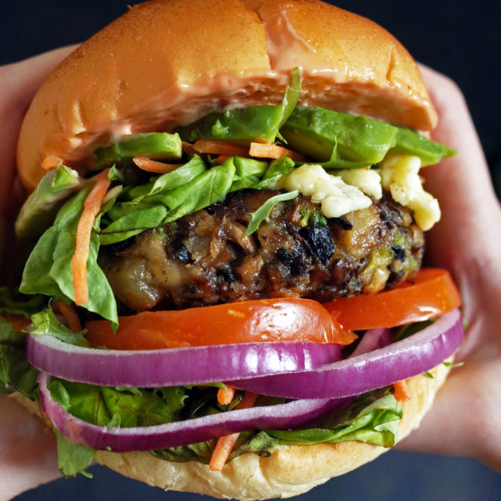 Hands holding a vegetarian mushroom burger within a brioche bun with salad, onions, tomatoes, cheese and avocado