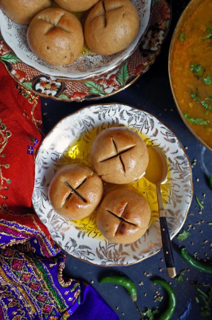 A plate of vegetarian baked bati covered in ghee on a dark background with a saree and whole green chillis