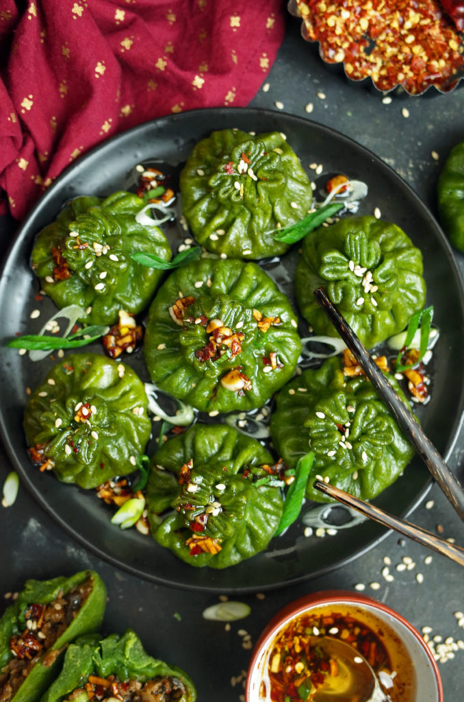 Spinach Dim Sum on a black plate topped with Chilli Oil, Sesame Seeds, and Spring Onion