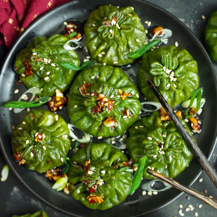 Spinach Dim Sum on a black plate topped with Chilli Oil, Sesame Seeds, and Spring Onion