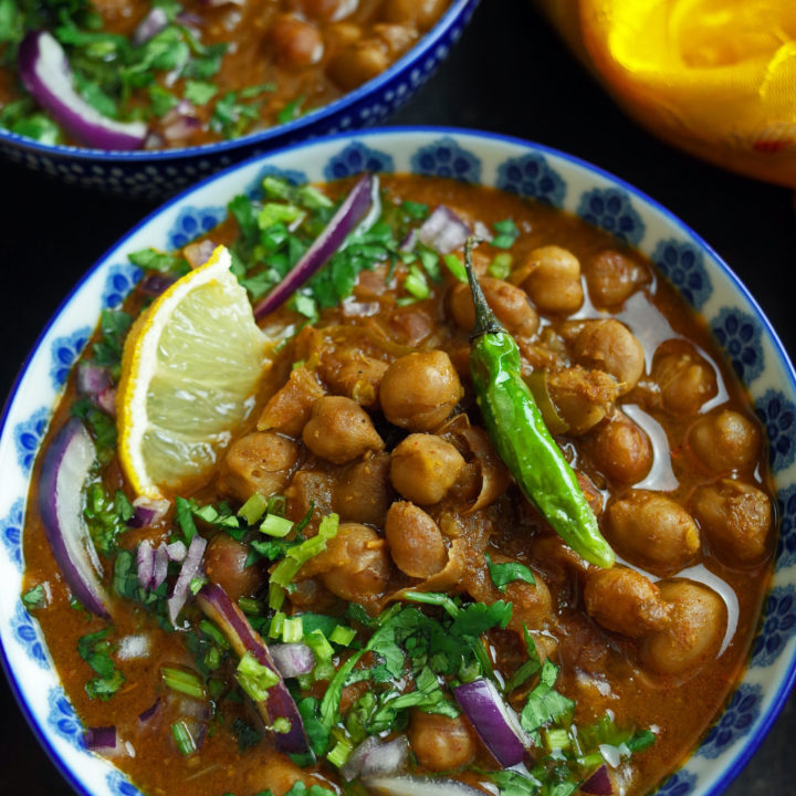 Two bowls of Vegan Chana Masala garnished with green chillis, coriander, onion and lemon slices