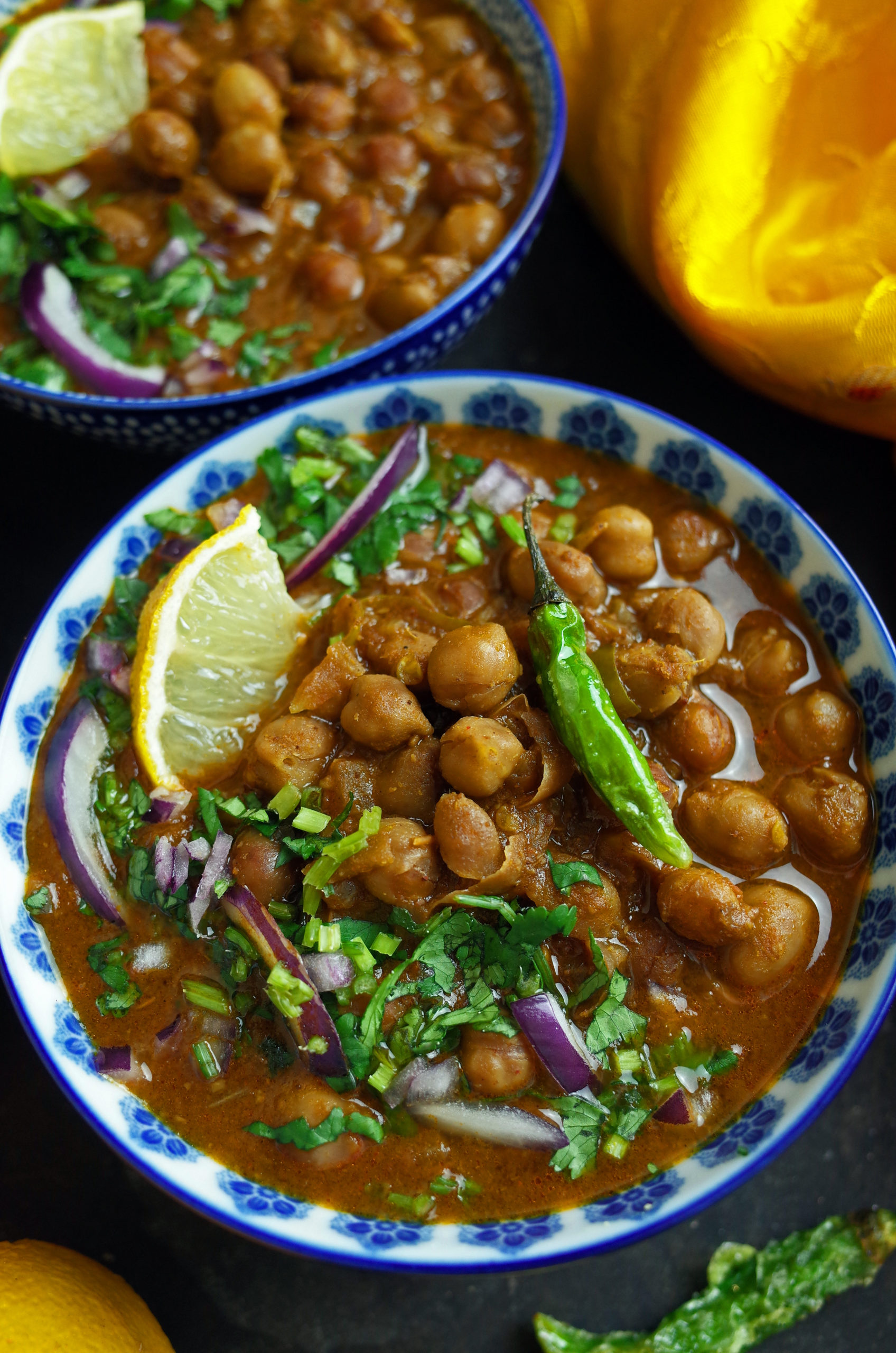 Two bowls of Vegan Chana Masala garnished with green chillis, coriander, onion and lemon slices