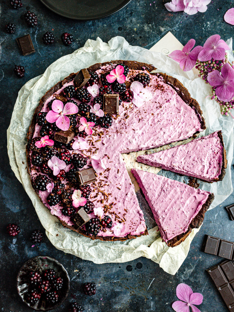 Pink-colored blackberry tart decorated with chocolate, fresh blackberries, and hydrangea flowers with two slices cut out, and more fresh blackberries in the background.