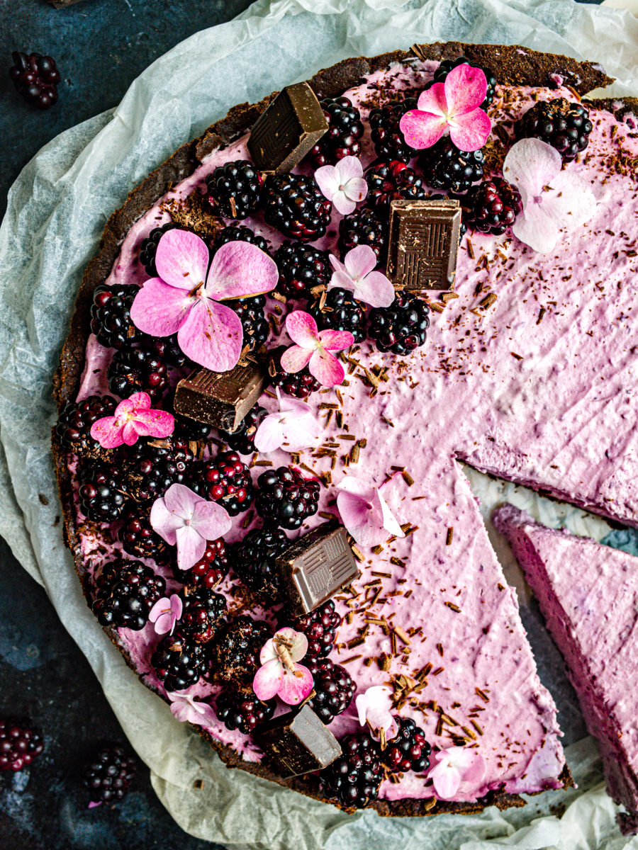 Close-up of a blackberry tart decorated with chocolate, fresh blackberries, and hydrangea flowers with a slice cut out, sitting on white parchment paper. 