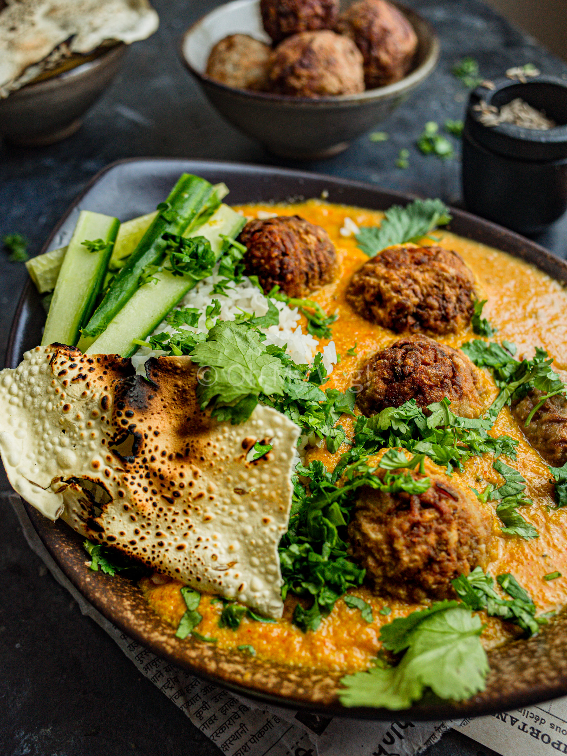 Vegan lauki kofta curry in an oblong brown bowl with rice, papad, and long cucumber slices. It's garnished with coriander. 