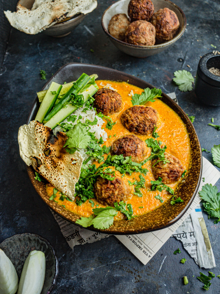 Vegan lauki kofta curry in an oblong brown bowl with rice, papad, and long cucumber slices. It's garnished with coriander. In the background are more kofta, papad, and onions.