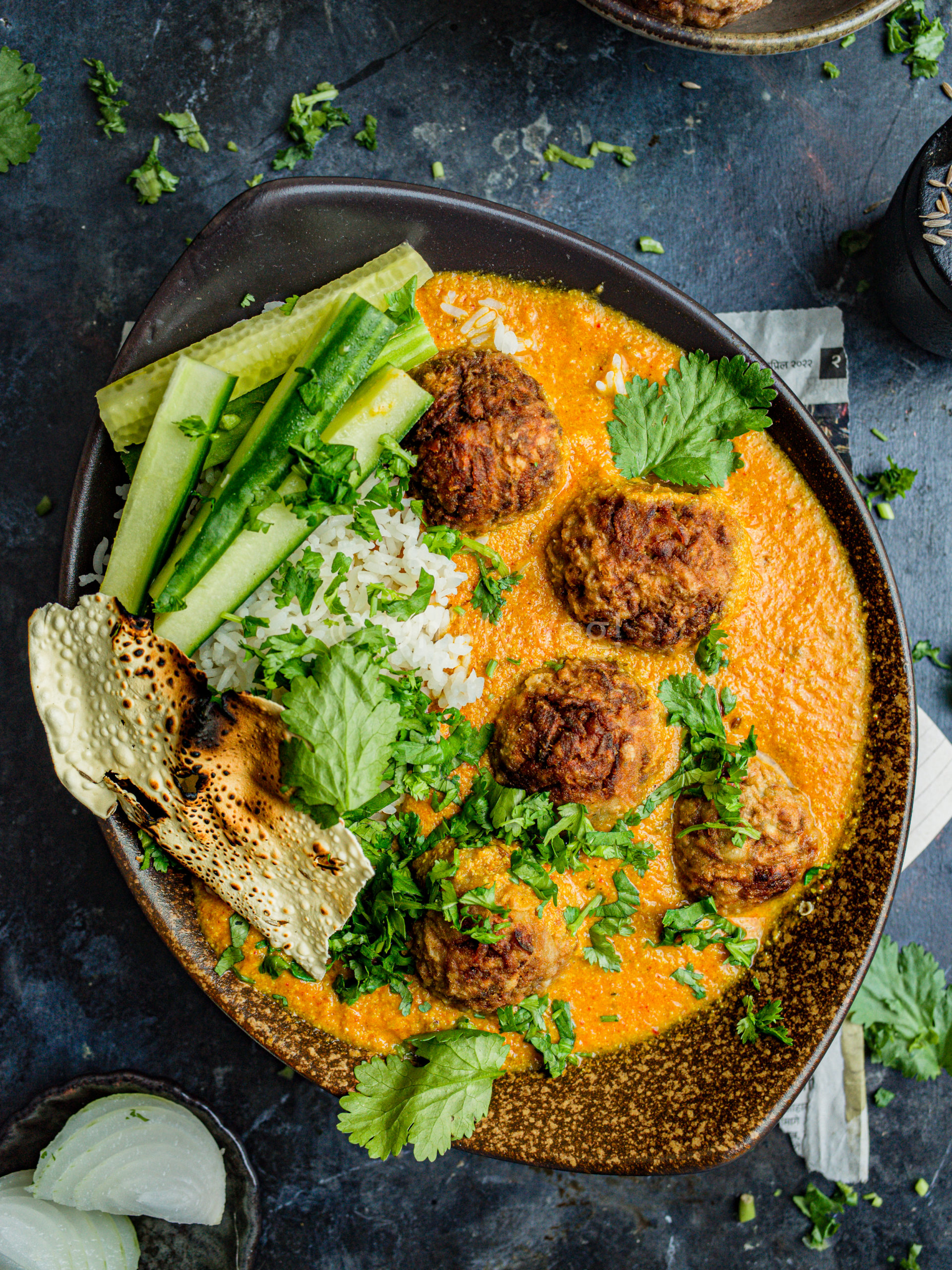 Vegan lauki kofta curry in an oblong brown bowl with rice, papad, and long cucumber slices. It's garnished with coriander.