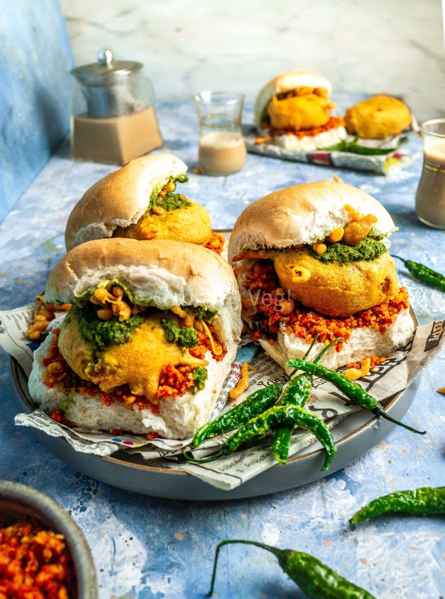 Three vada pav on a plate covered in newspaper, along with fried green chilis and tea in the background.