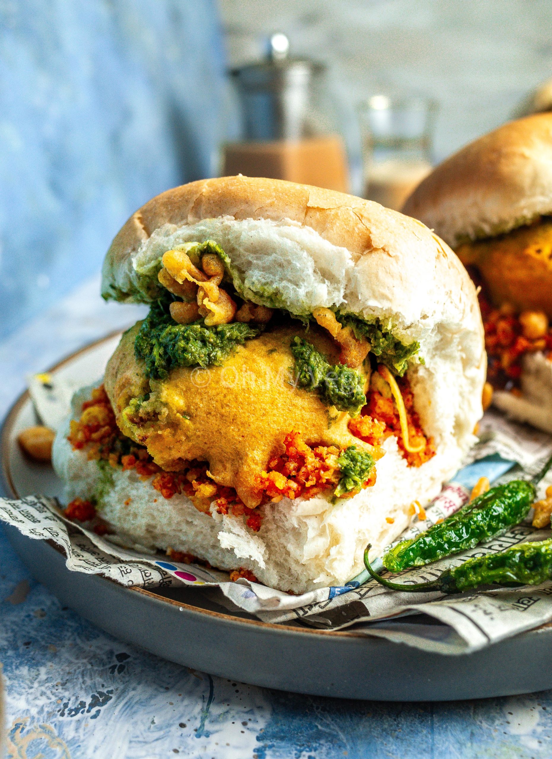 Vada pav on a blue background alongside fried green chilis.