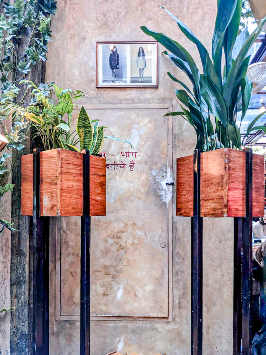 Detail of the interior of Dishoom Carnaby, featuring two large standing plant pots, with a background of a beige lime washed wall, red Hindi text painted on the wall, and two vintage Indian photographs. 