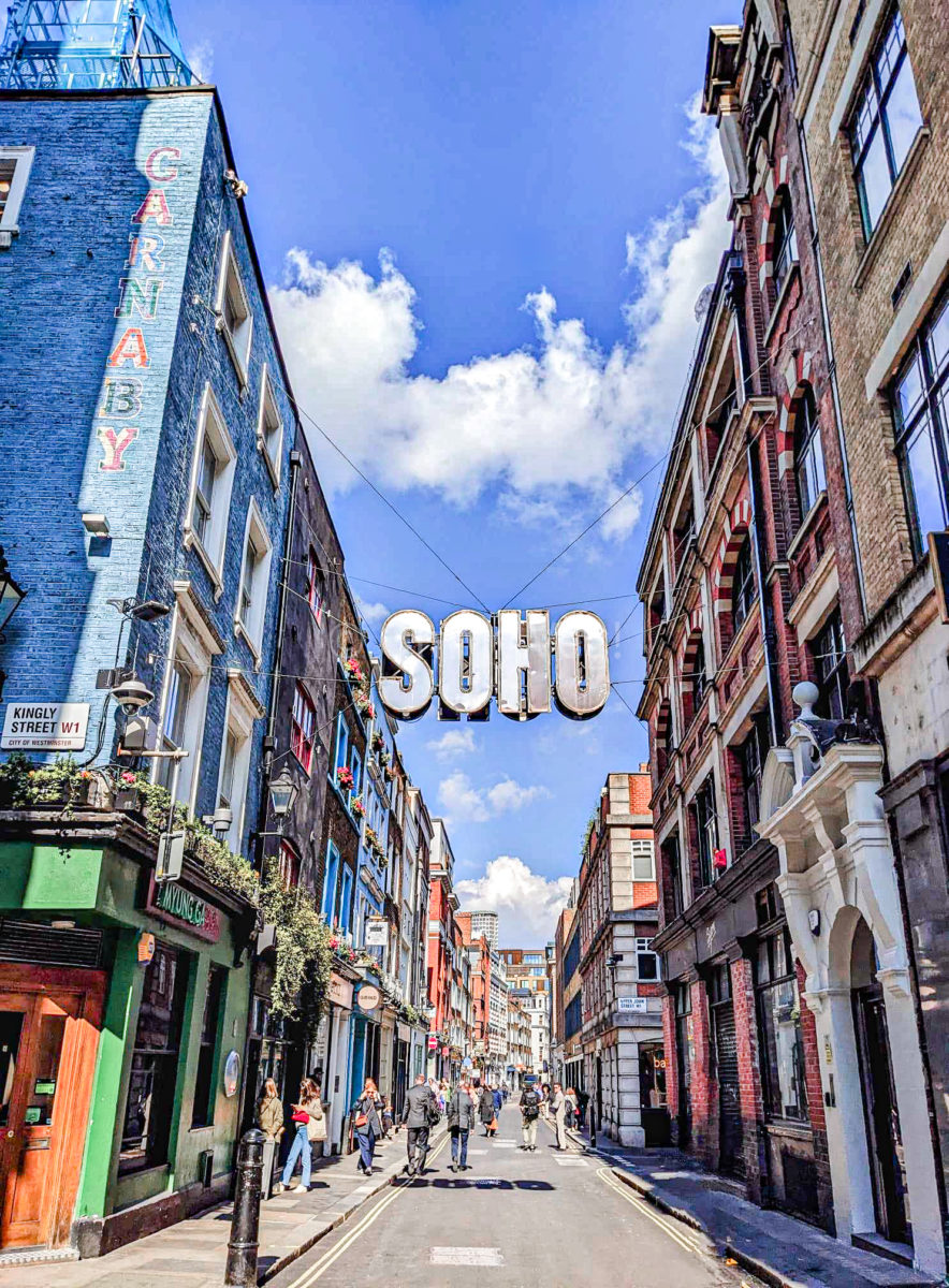 A London street with blue skies, and a large silver sign saying "SOHO" hanging between the buildings. 
