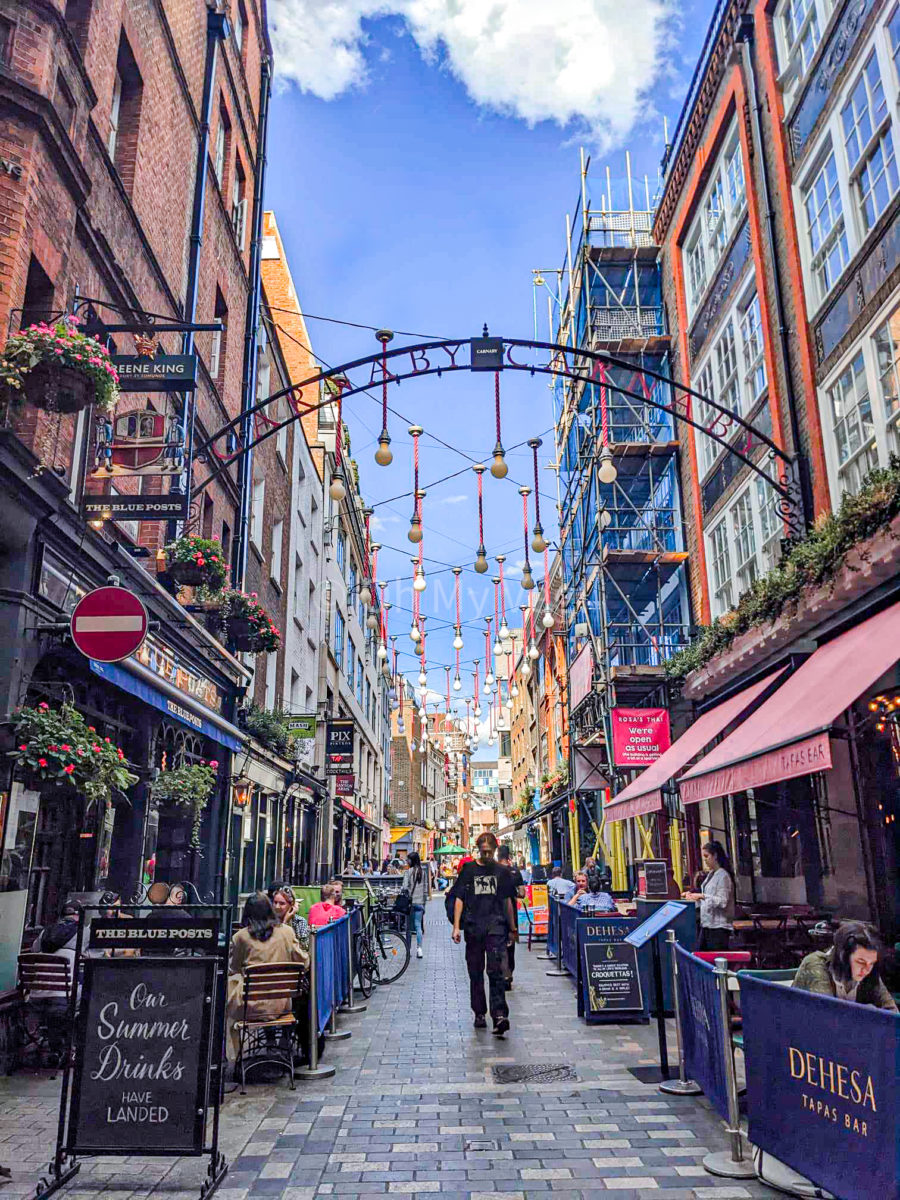 A busy London street with a blue sky. There is a metal arch saying "Carnaby, Carnaby" hanging between the buildings, and lights strung all along the street, which is filled with cafes. 