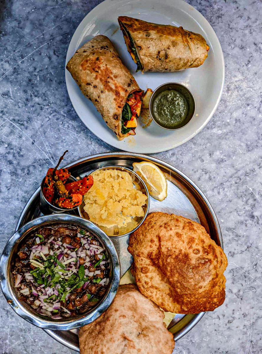 A thali with chole, carrot pickles, sooji halwa, puri, and a slice of lemon on a marble table top with paneer rolls and mint chutney on another white plate. 