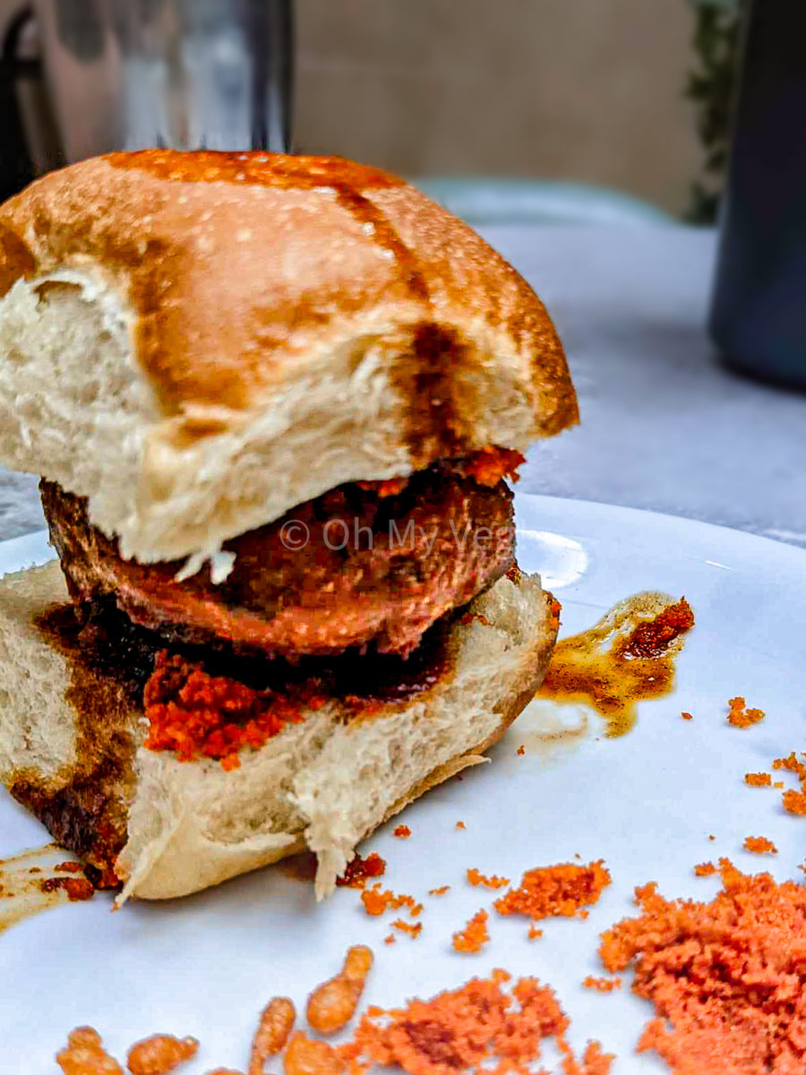 Vada pav on a white plate, with batata vada in the bun, tamarind chutney, and dry garlic chutney. 