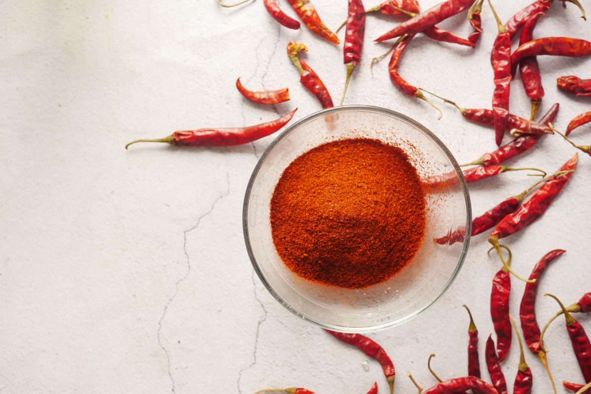 Red chili powder in a small glass bowl with whole red chilis in the background. 