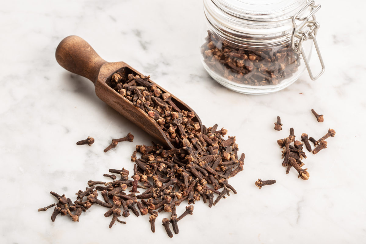 A small wooden spoon holding cloves on a marble background, with a jar of more cloves in the distance. 