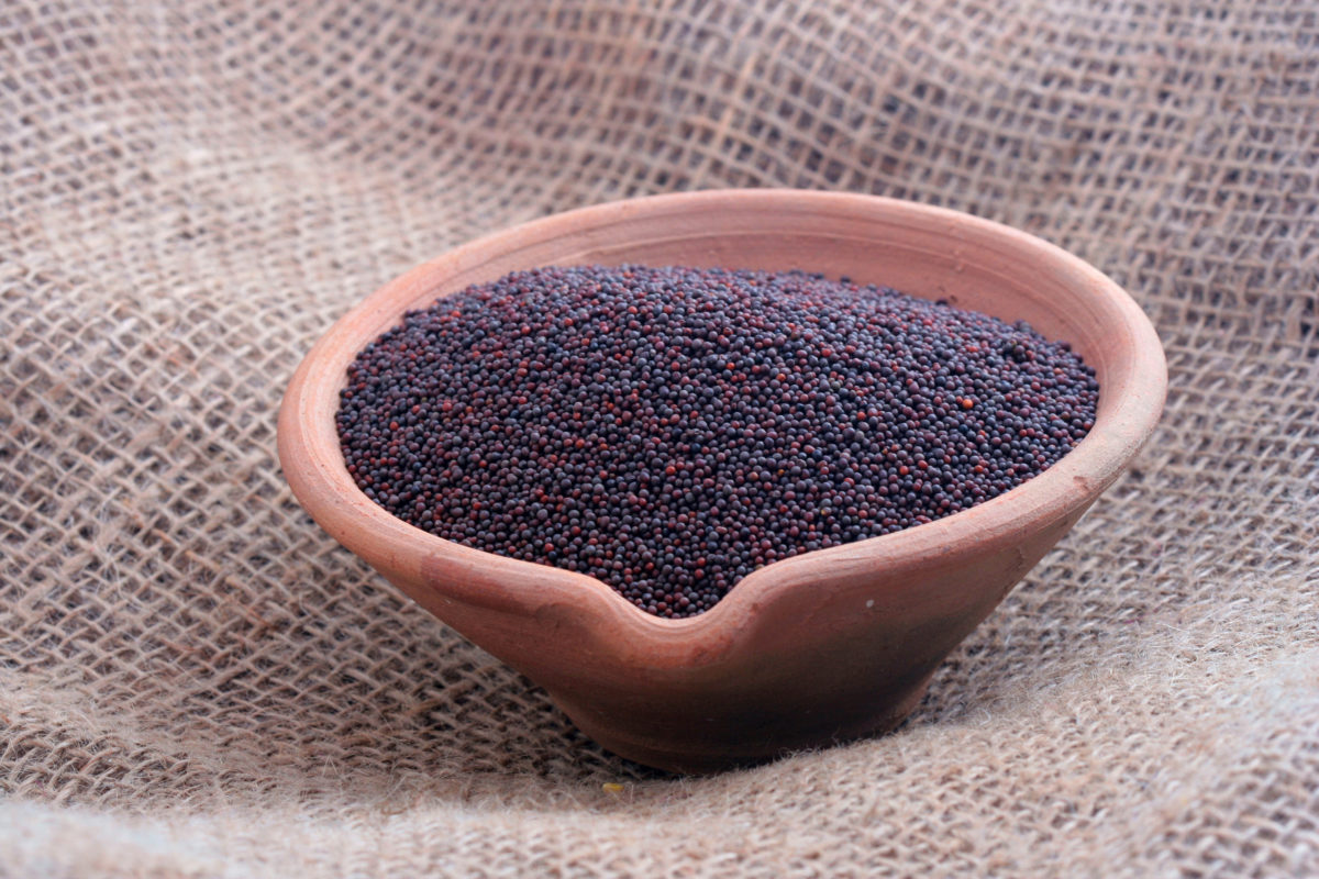 A small clay bowl full of black mustard seeds on a woven brown cloth. 