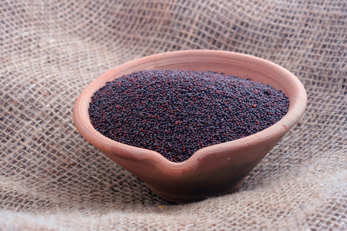 A small clay bowl full of black mustard seeds on a woven brown cloth.