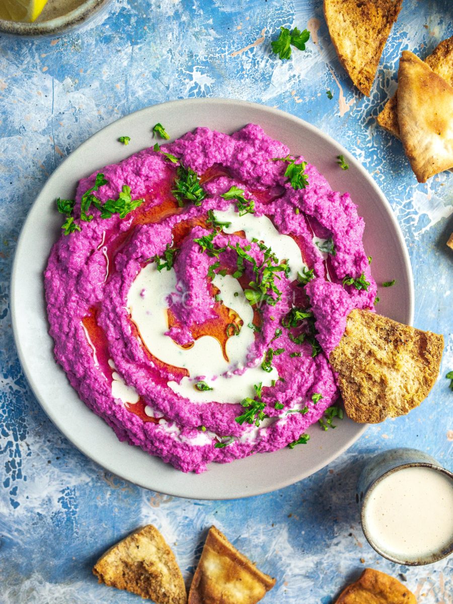 A plate of beetroot hummus topped with parsley and garlic tahini sauce, on a blue background with scattered pita chips. 