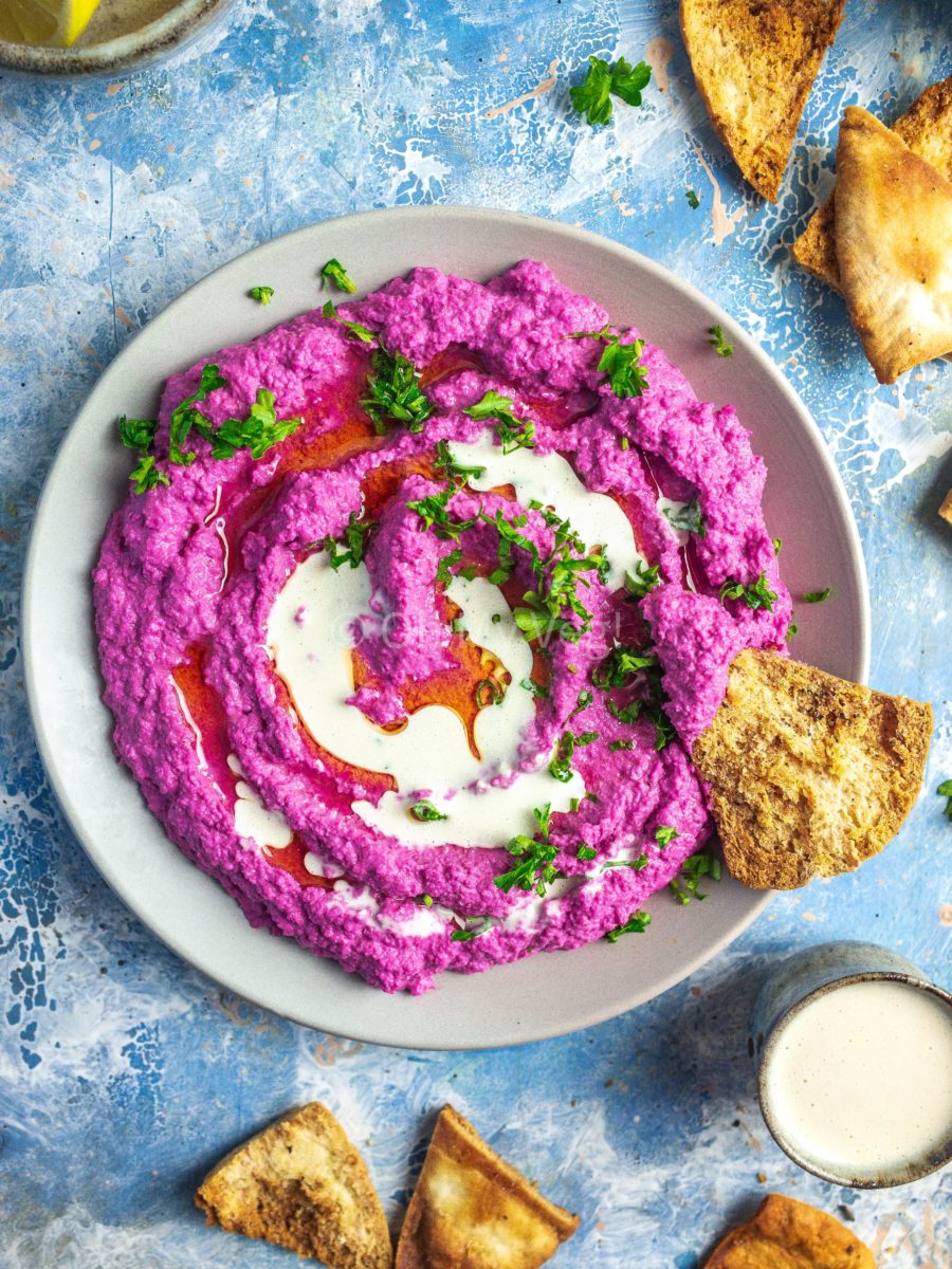 A plate of beetroot hummus topped with parsley and garlic tahini sauce, on a blue background with scattered pita chips.