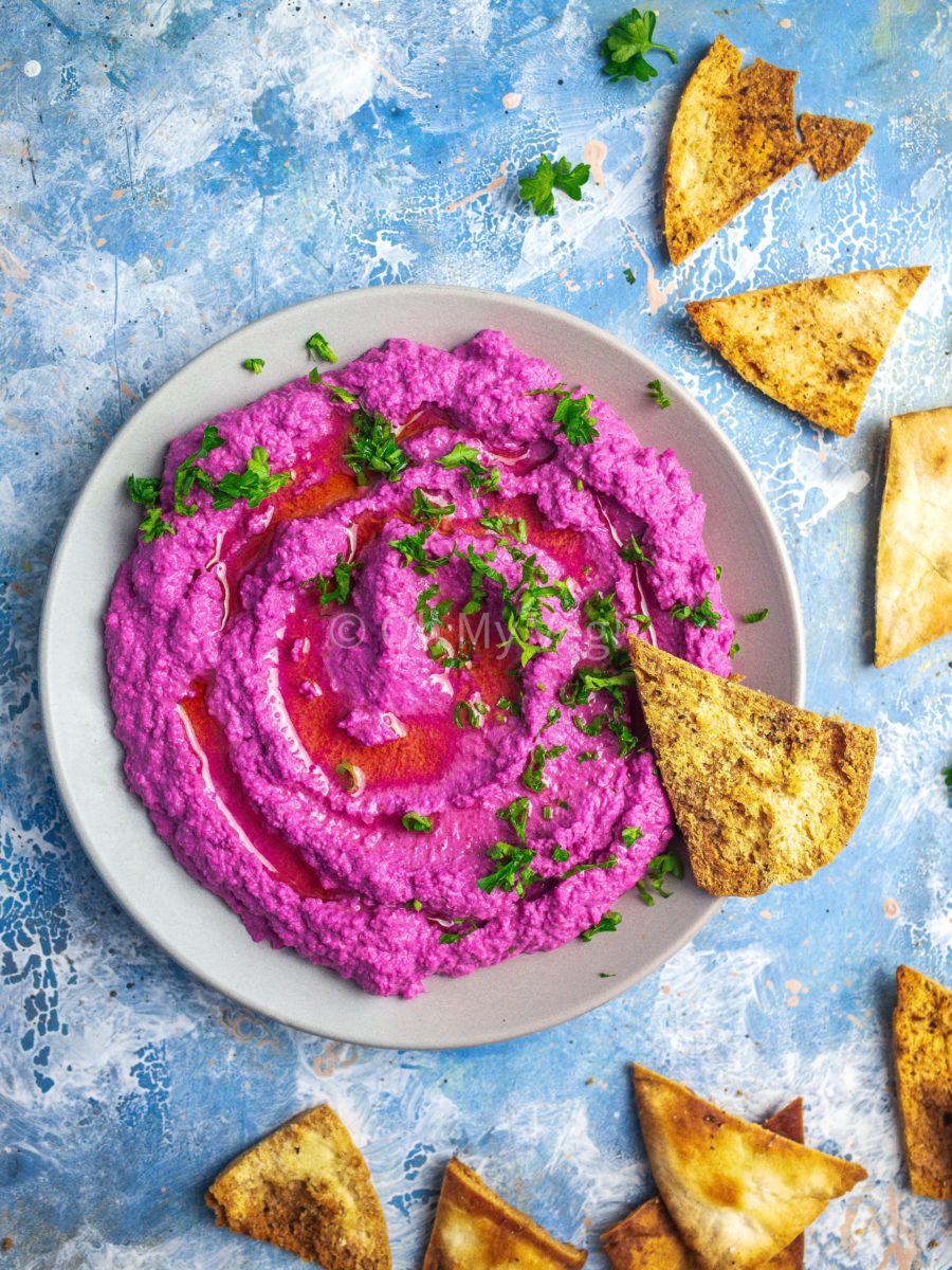 A plate of beetroot hummus topped with parsley, on a blue background with scattered pita chips. 