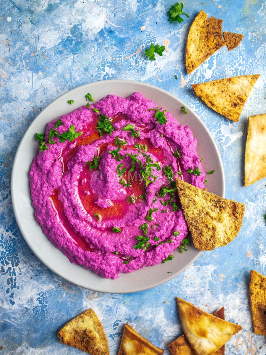 A plate of beetroot hummus topped with parsley, on a blue background with scattered pita chips.