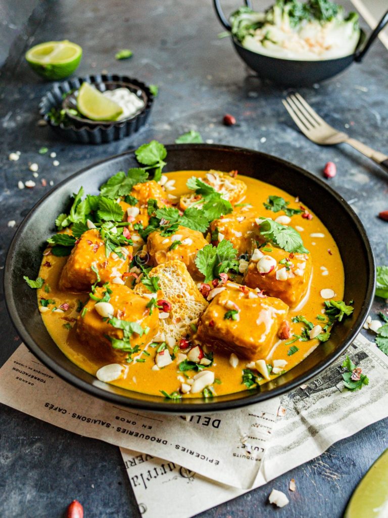 Tofu satay curry in a black bowl, with garnishes and a small bowl of pak choy in the background.