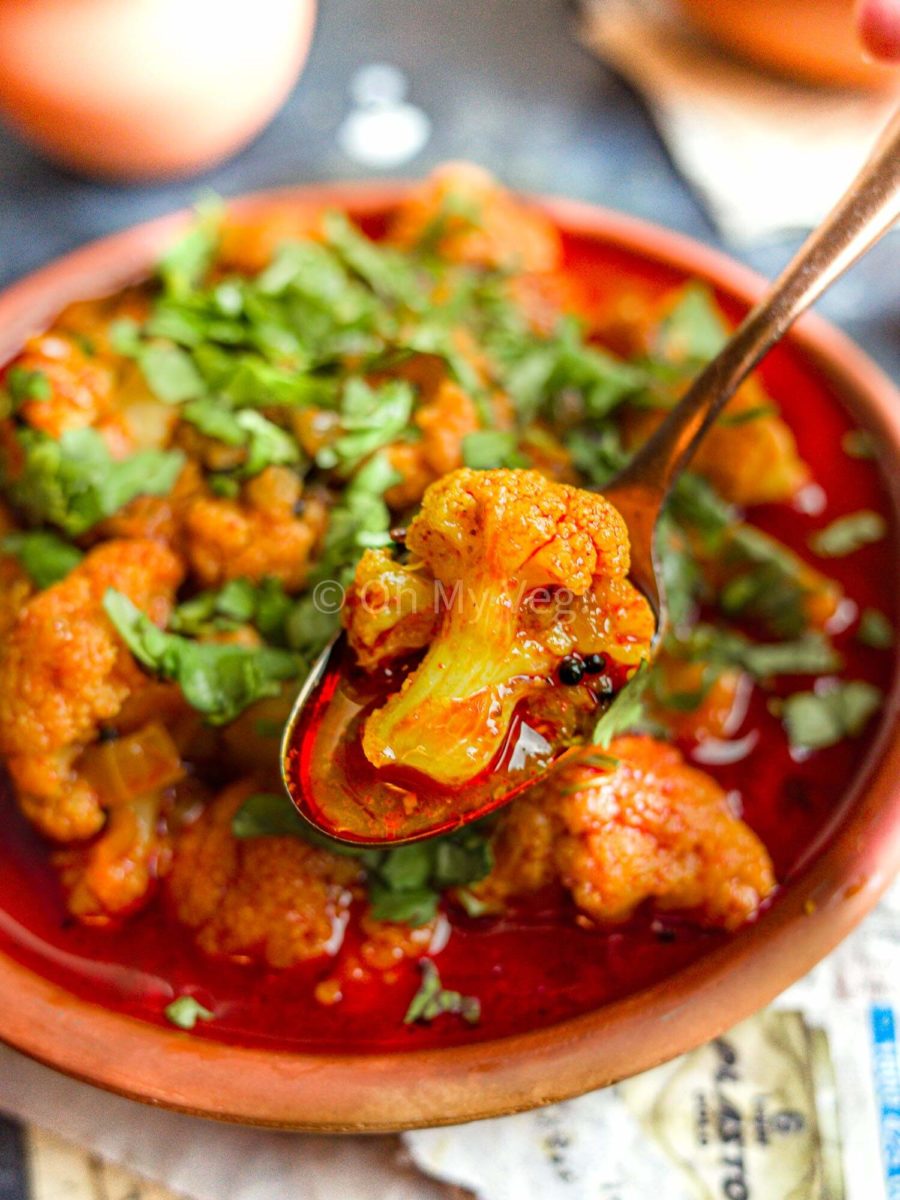 A spoon with a piece of curried cauliflower hovering above a clay bowl full of Maharashtrian Flower Batata Rassa. 