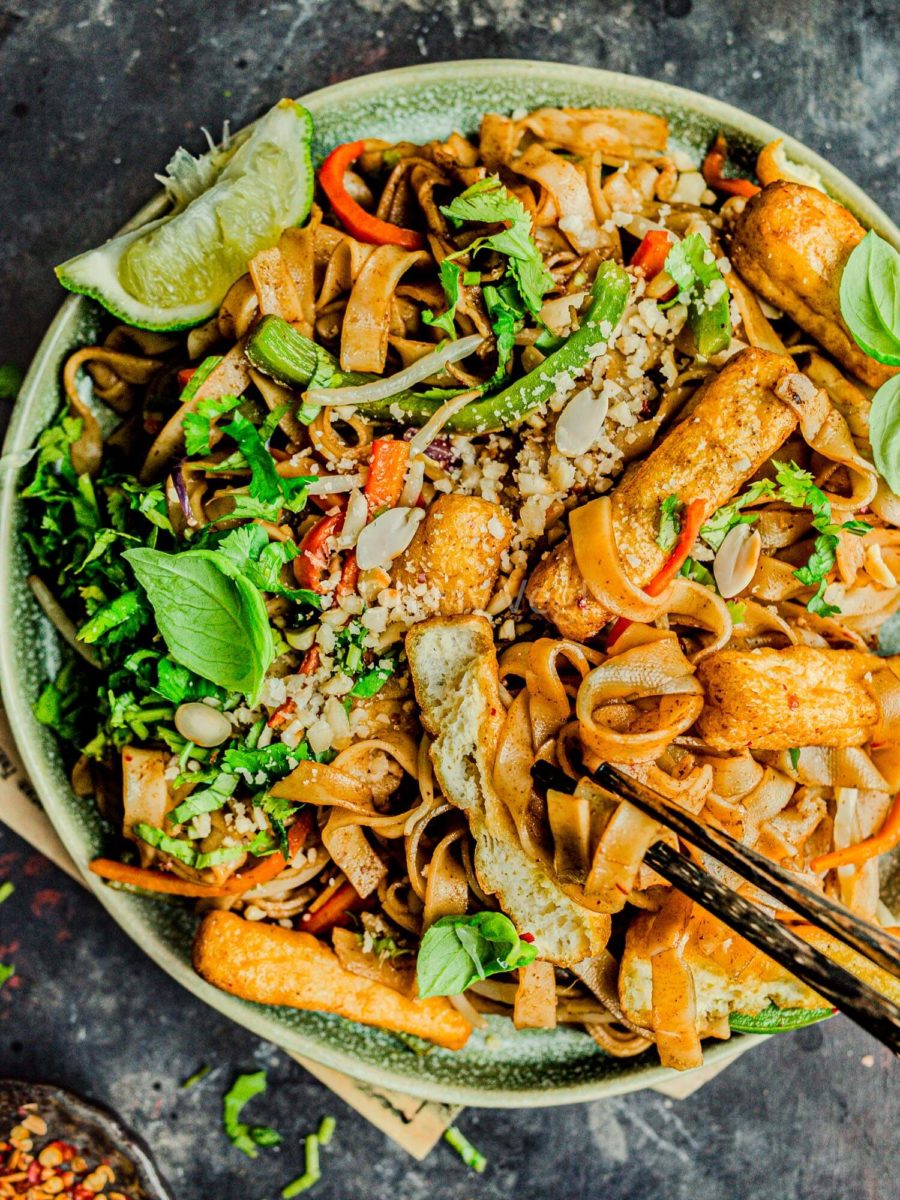 A plate of vegan pad thai with tofu and vegetables on a plate, with chopsticks picking up the noodles.