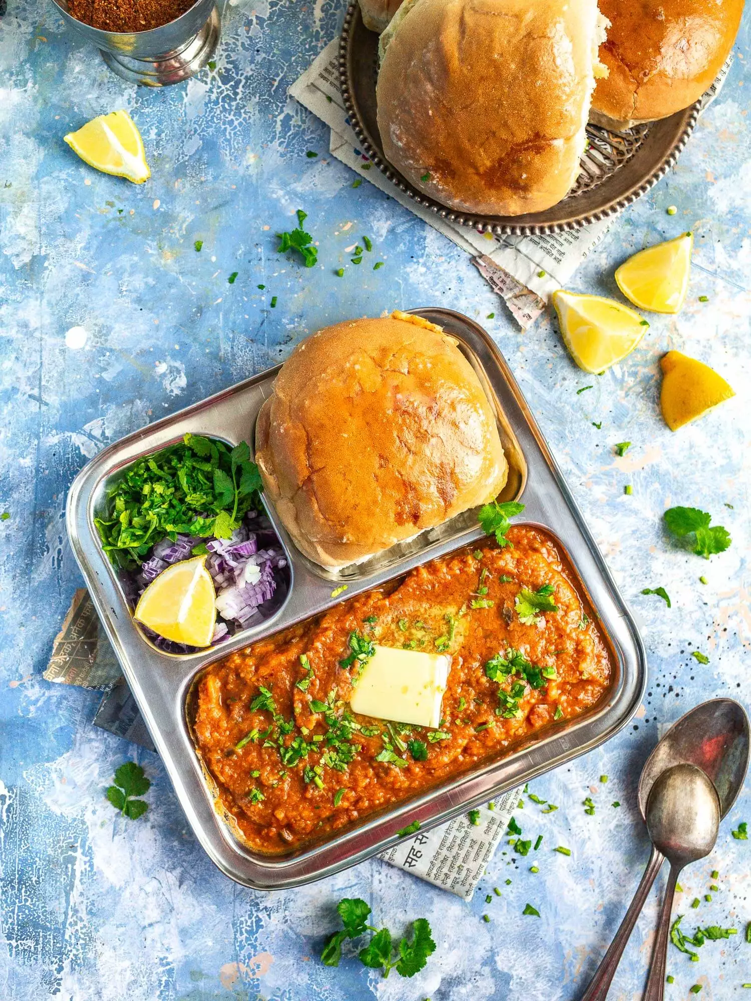Pav bhaji in a metal tray on a blue background, with pav in the background.