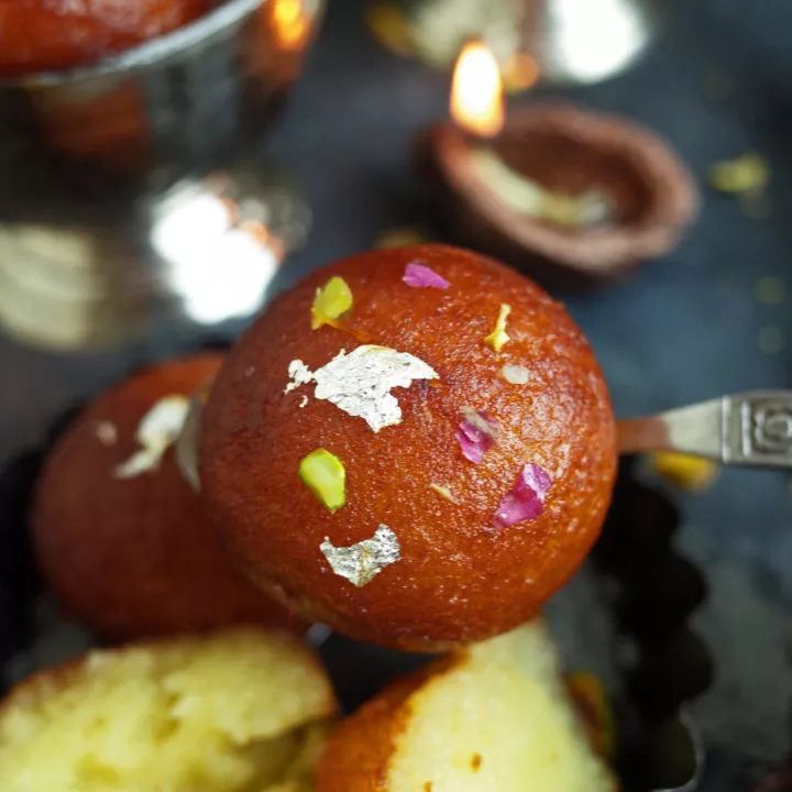 A Gulab Jamun topped with rose petals, pistachios and silver leaf being held on a spoon, with more Gulab Jamun in the background