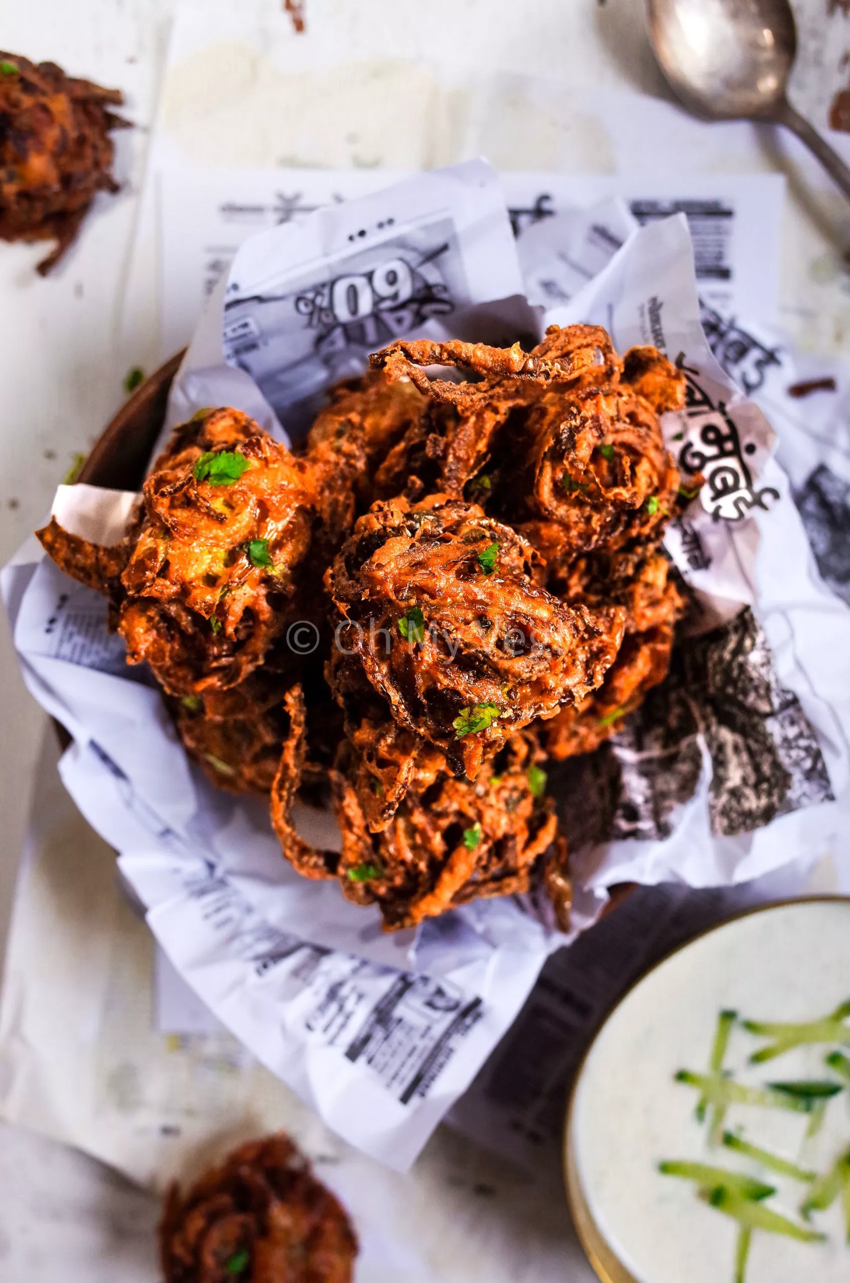 A pile of onion bhajis on newspaper with yoghurt raita on the side