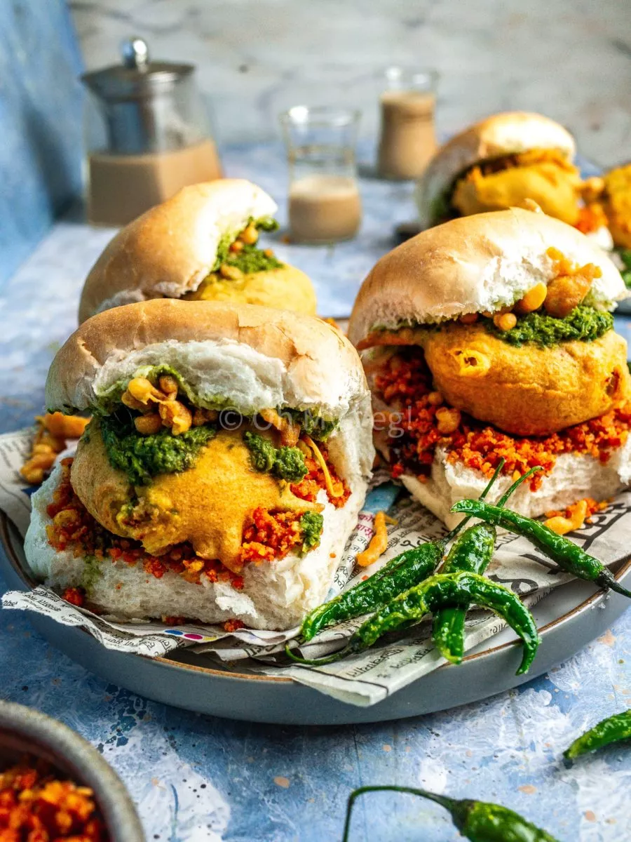 Vada pav on a plate covered in newspaper with fried green chilis, on a blue background.