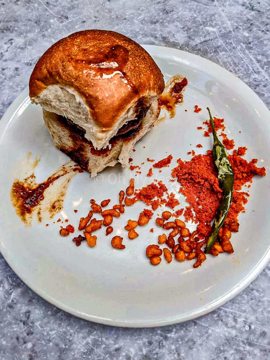 Vada pav on a white plate with a fried green chili, red dry garlic chutney, and a smear of tamarind sauce. 