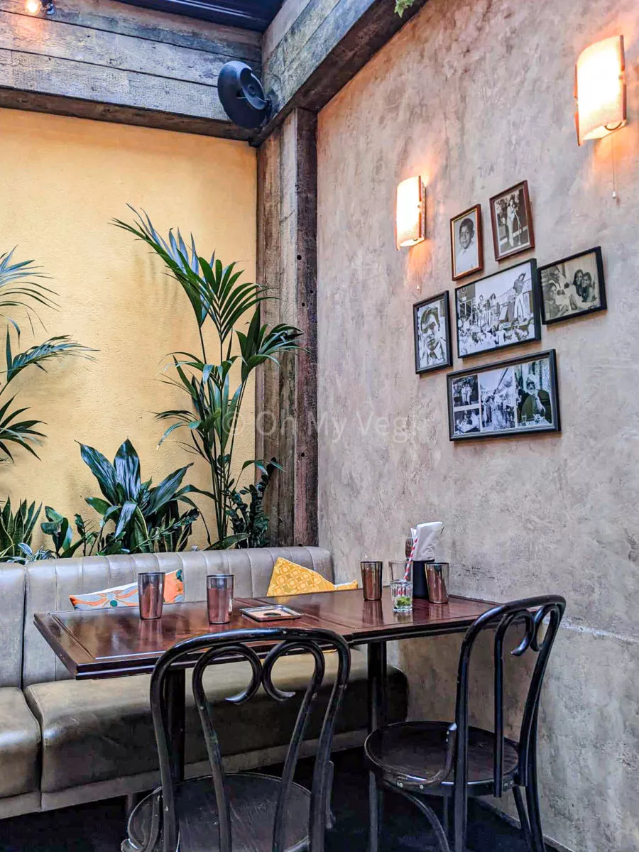 Interior of Dishoom Carnaby, with a table, one yellow wall, green foliage, and a lime washed grey wall with vintage photos. 
