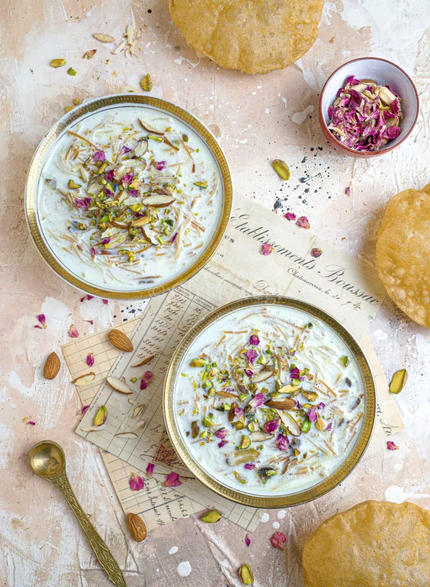 Two small bowls of seviyan kheer on a beige background, with scattered nuts, rose petals, and puris. 