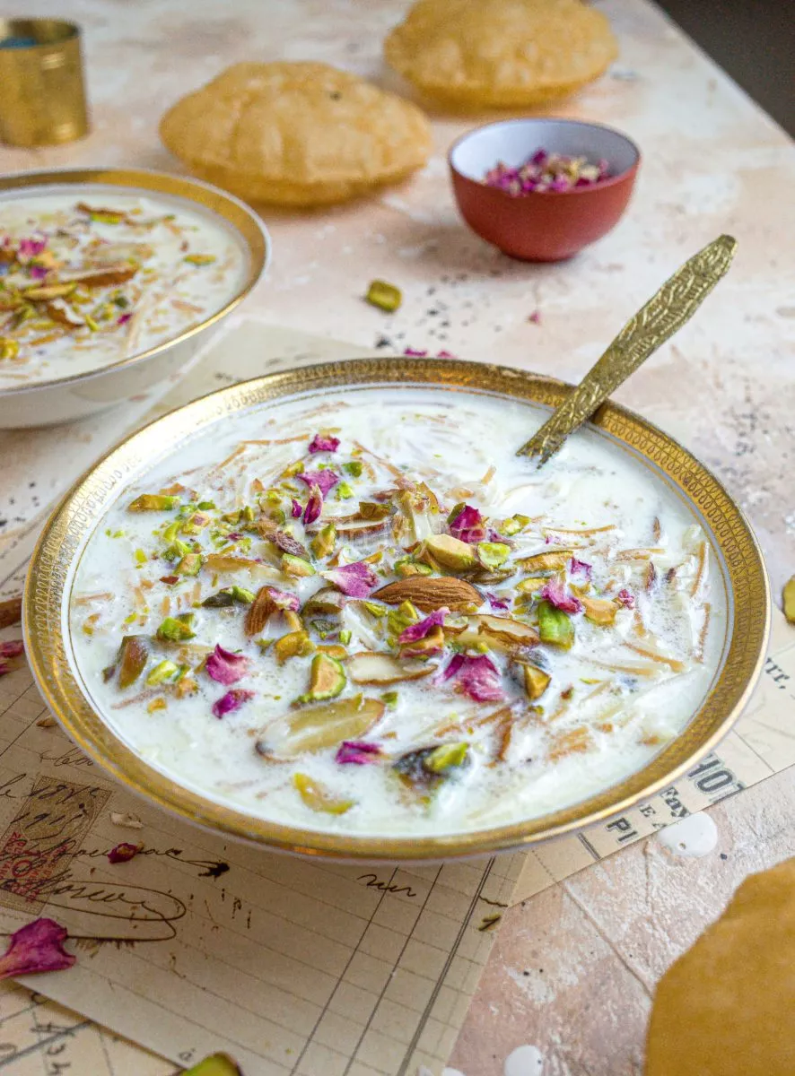 Seviyan kheer in a small golden bowl, topped with nuts and rose water, with a gold spoon. There are puris in the background. 