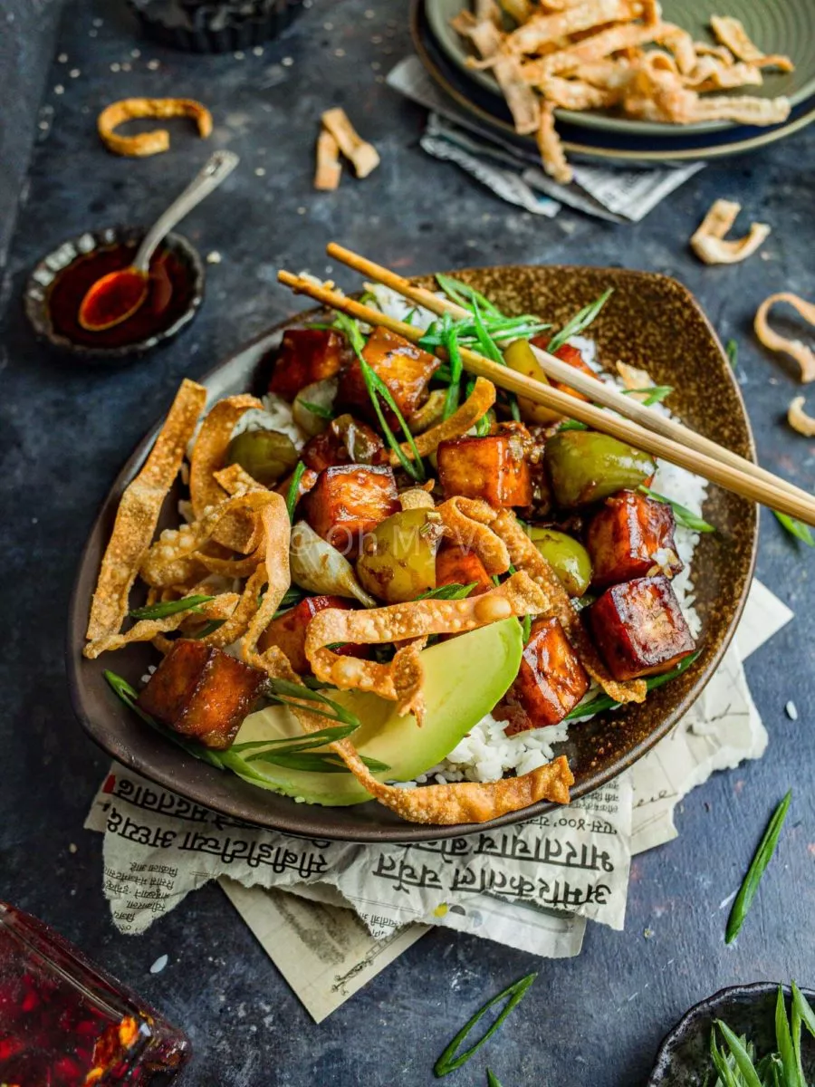 Chilli paneer in a bowl with rice, avocado, and wonton crisps, on a dark blue backdrop.