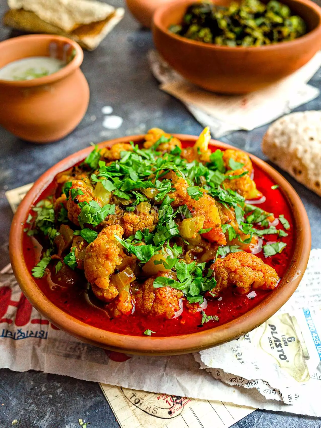 Maharashtrian Flower Batata Rassa in a clay pot, with raita and bhindi masala in the background.