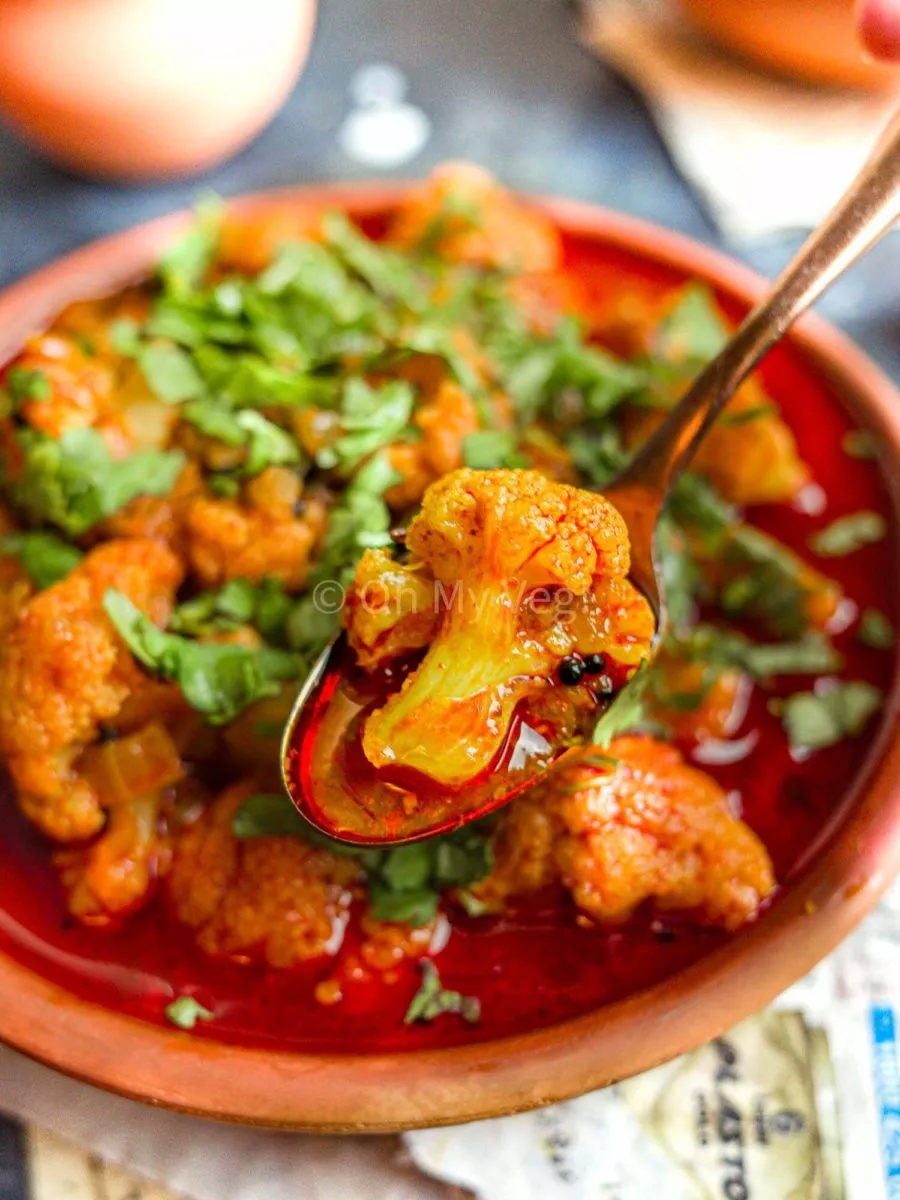 A spoon with a piece of curried cauliflower hovering above a clay bowl full of Maharashtrian Flower Batata Rassa.