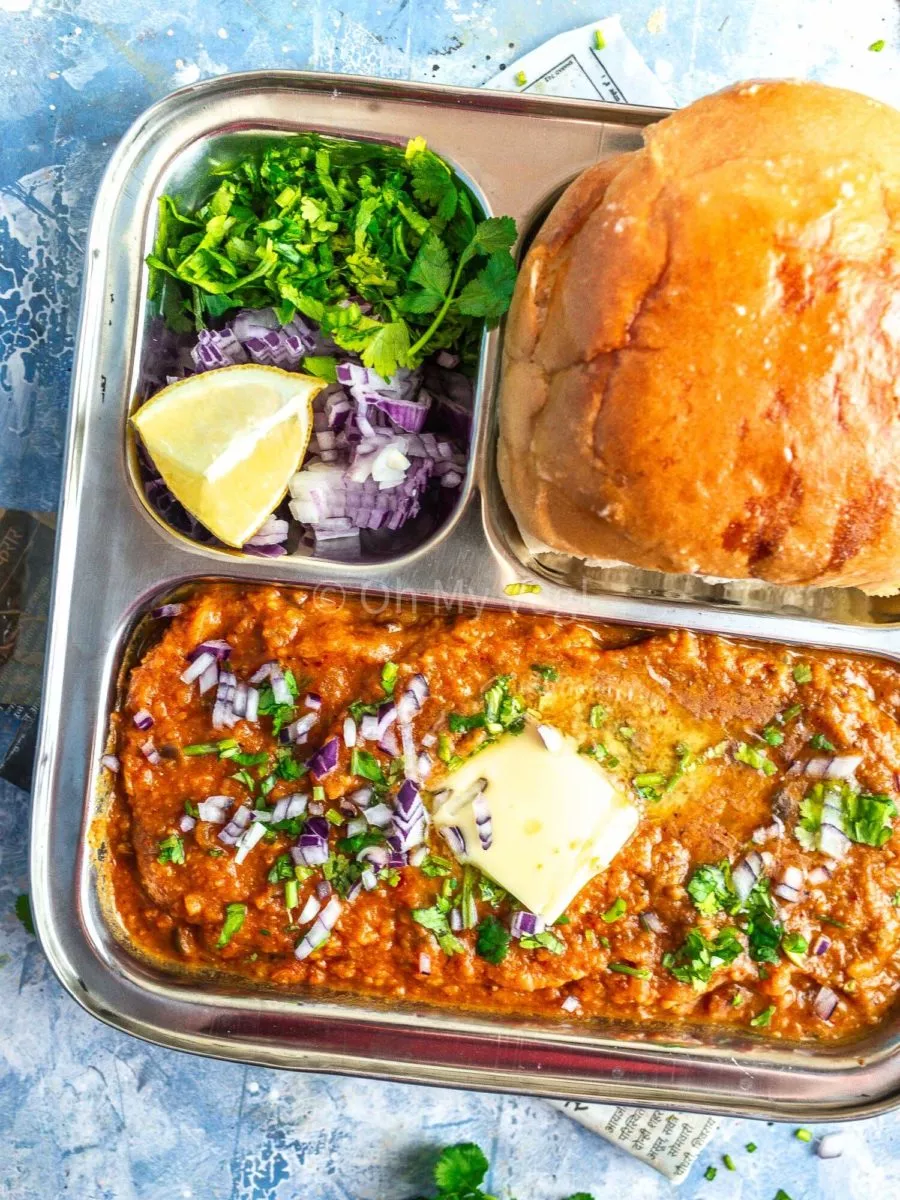 Mumbai street style pav bhaji in a metal tray with butter and garnishes on top. 