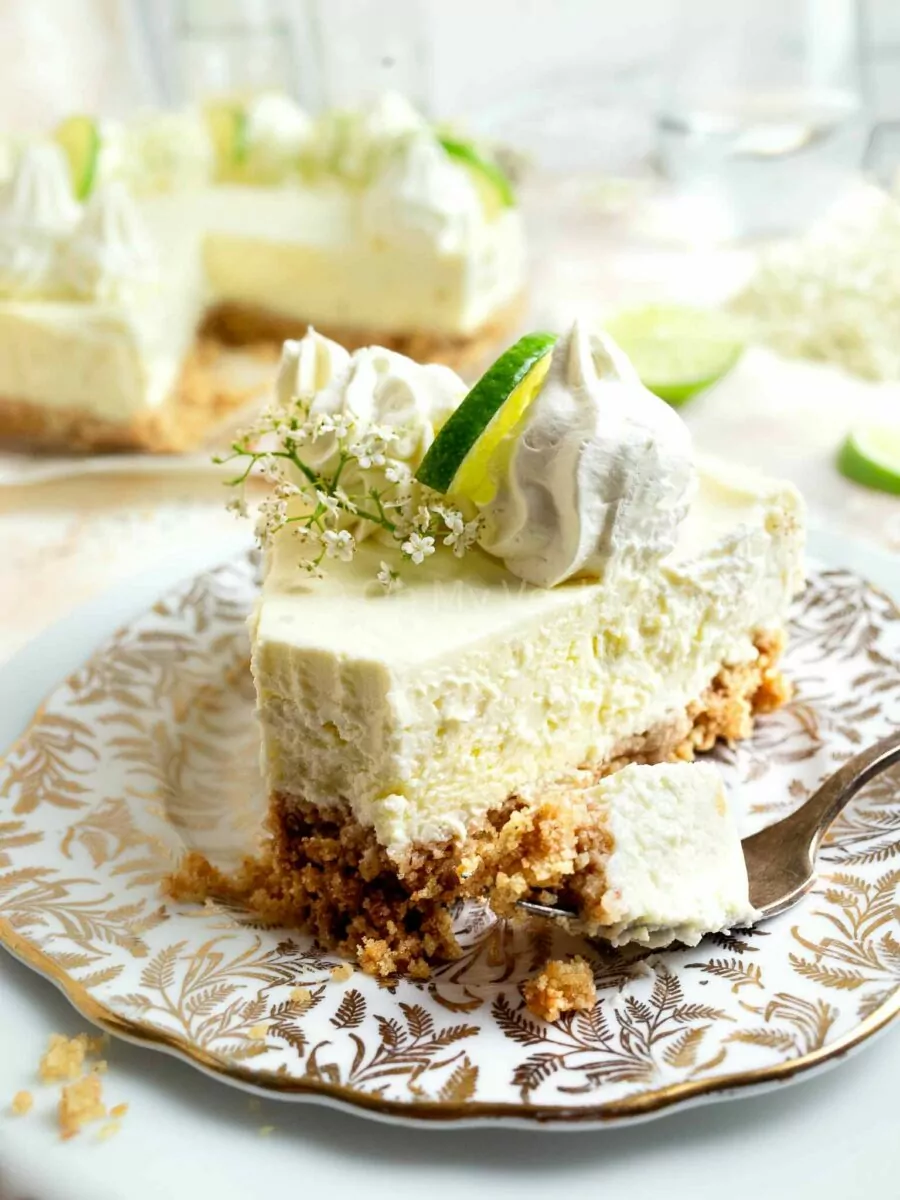 A piece of elderflower cheesecake with a fork, decorated with whipped cream and limes. 